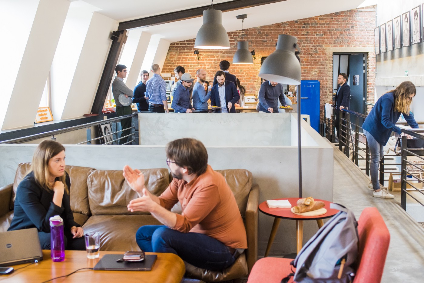 Photo of a bright, open-style office with two people conversing on a couch in the foreground and multiple people standing and conversing in the background.