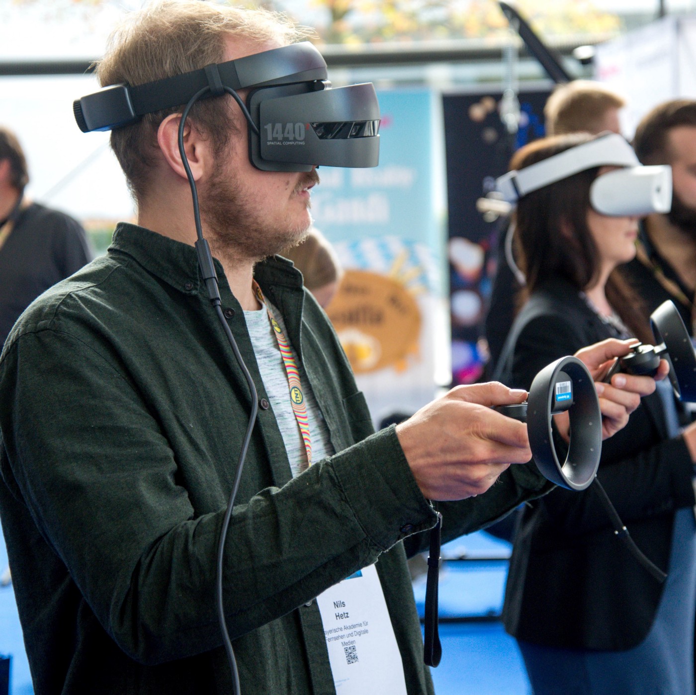Bearded man with a virtual reality headset on, holding controllers in each hand with others in the backround also using virtual reality systems.
