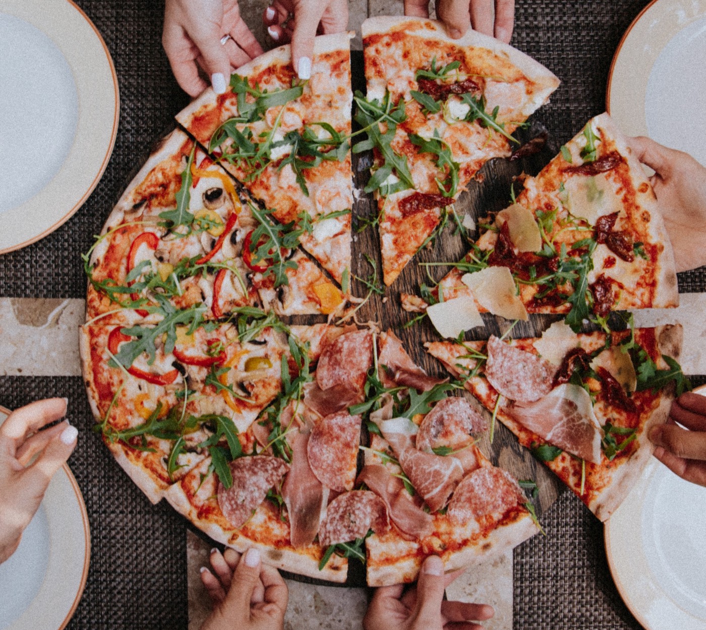 A pizza, half vegetables and half meat, sliced into eight pieces with eight hands each gripping a slice. The pizza is surrounded by small white plates.